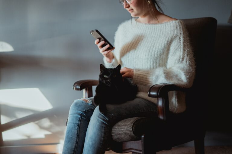 Frau sitz auf einem Sessel und schaut auf Ihr Smartphone auf ihrem Schoß liegt eine Katze, die von ihr gestreichelt wird