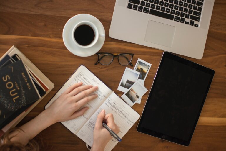 Aufsicht auf einen Tisch mit Arbeitsmaterialien wie Laptop Tablet Notizbuch und einer Tasse Kaffee, eine weibliche Hand schreibt etwas in einen Kalender
