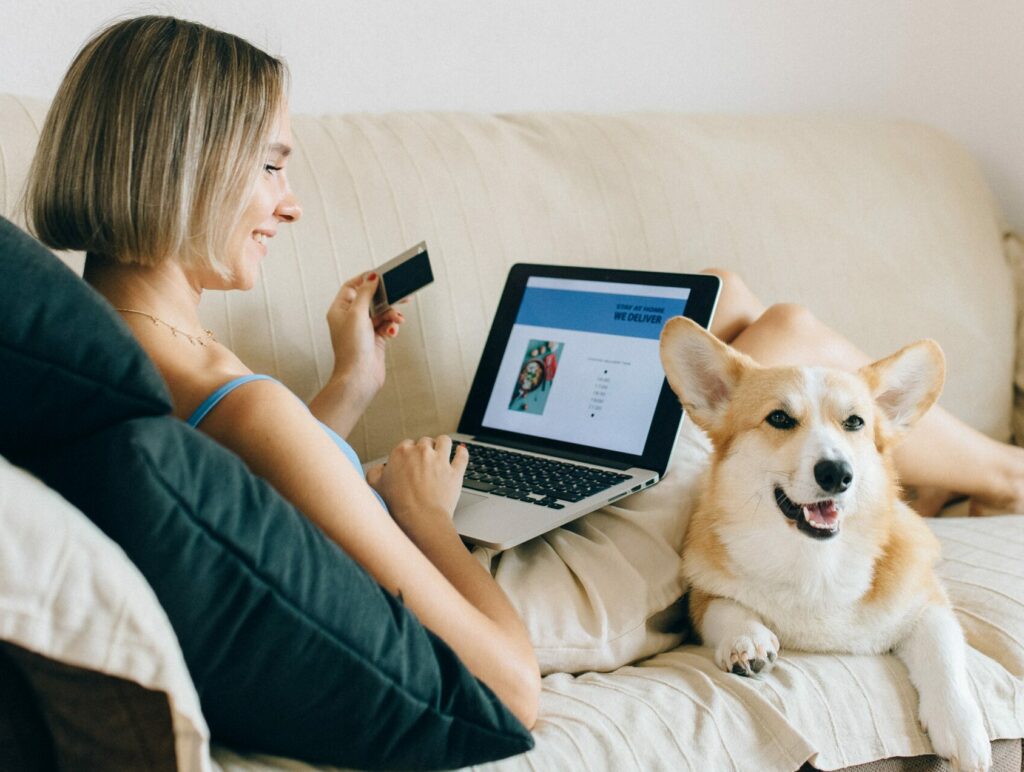 Frau liegt auf dem Sofa mit Laptop auf dem Schoß, sie lächelt, daneben liegt ihr Hund, digitale Tierarztpraxis